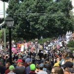 A large crowd of anti-racist protestors face off against white supremacists on a tree-lined street. Only the tops of most people's heads are visible. there are two neon hats in the crowd. They are National Lawyers Guild legal observers.