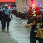 A group of police officers in blue shirts approach down a wet street with guns pointed toward protestors who duck their heads and run away from them. There are cop cars with lights flashing in the background.