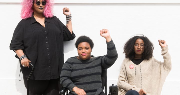 Torso level photo of three Black and disabled folx (a non-binary person holding a cane, a woman in a power wheelchair, and a woman on a folding chair) raising their fists on the sidewalk in front of a white wall. Credit: Disabled and Here