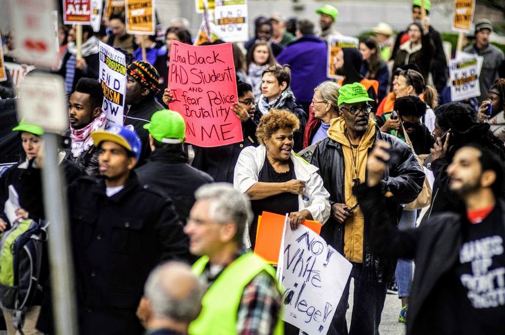 In a crowd of anti-racist activists holding signs in support of Ferguson and denouncing police brutality, there are 5 folks wearing neon green hats. One of them, a black man with glasses, faces the camera. He is NLG Legal Observer Blair Anderson.